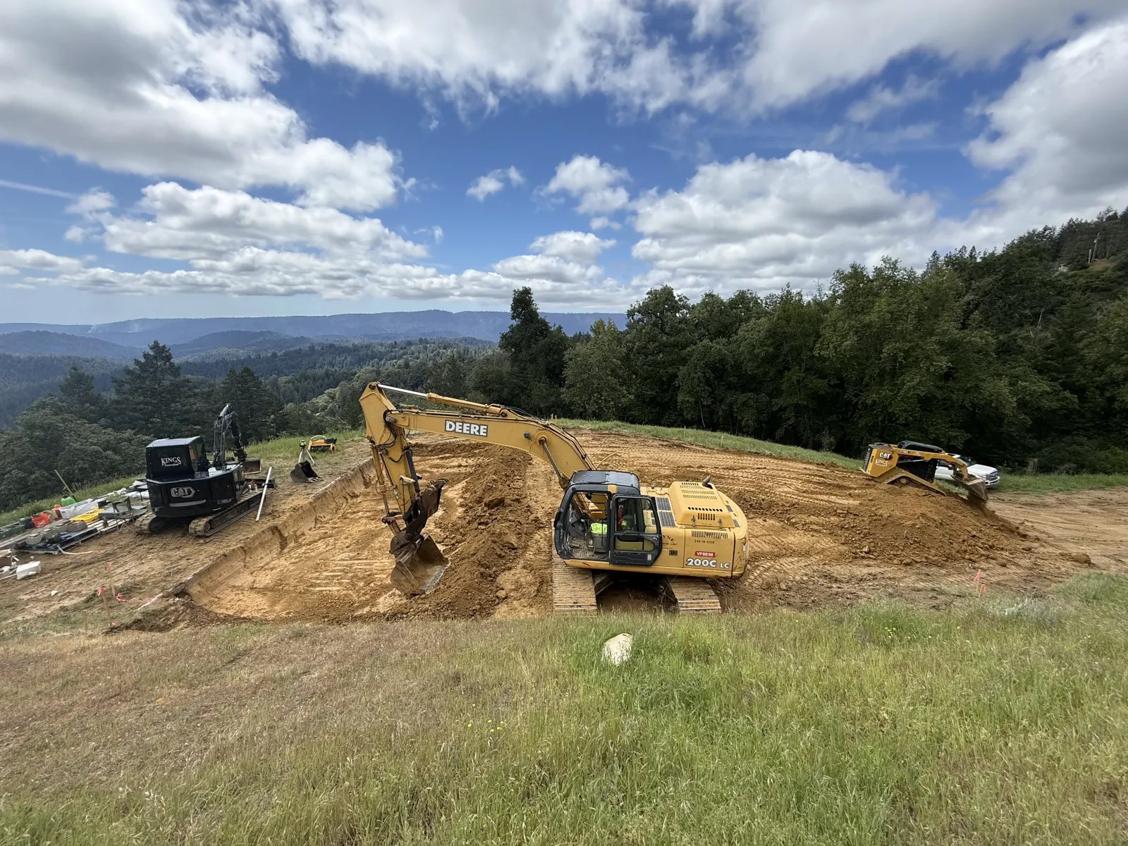 Excavator and dozer cutting a hilltop building pad with forested ridges beyond