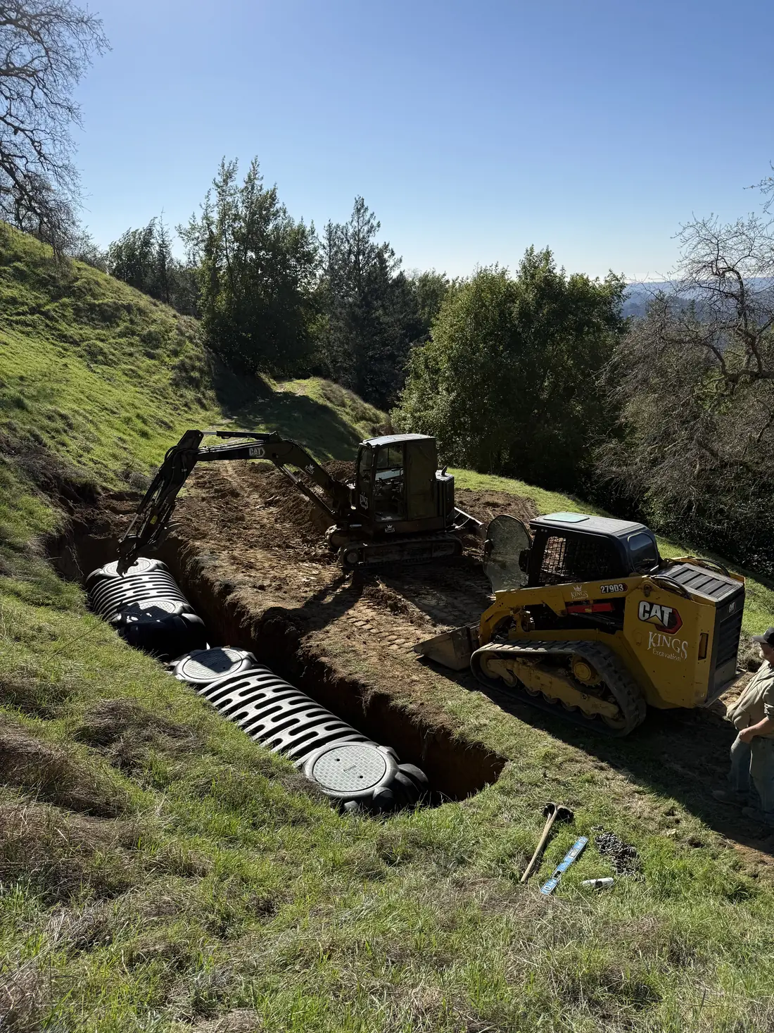 Skid steer setting a septic tank in a green meadow with a large oak